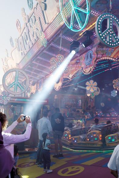 Ein Fahrgeschäft auf einem Jahrmarkt mit leuchtenden Friedenszeichen und Blumenmotiven, die mit bunten Lichtern geschmückt sind. Ein heller Scheinwerferstrahl fällt auf die Szene, in der mehrere Personen stehen, darunter ein Kind und Erwachsene. Eine Person im Vordergrund hält eine Kamera oder ein Smartphone, um ein Foto zu machen.