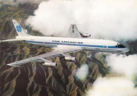 A Pan American World Airways Douglas DC-8 soars above a rugged mountain landscape partly veiled by drifting clouds. The aircraft wears Pan Am’s iconic white and light-blue livery, with the globe logo on the tail and “PAN AMERICAN” written boldly along the fuselage. Four silver jet engines hang beneath the swept wings, catching soft light as the plane cuts through the sky. Below, steep ridges and shadowed valleys form a dramatic pattern, while patches of clouds rise around the aircraft, giving the impression of effortless altitude and classic jet-age elegance. The scene evokes the pioneering spirit of early long-range commercial flight and Pan Am’s role as a symbol of international travel.