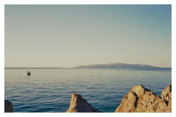 Color photograph of the Adriatic Sea with a view of the island of Cres, taken from a bathing spot between Ika and Ičići. Although taken in the morning, the light is already very bright. The small rocks in the foreground glow bright gold, and the sky ranges from turquoise to almost white on the horizon. A small boat floats in the water.