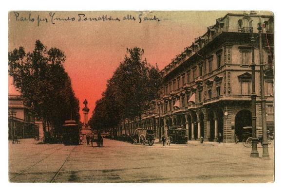 The image depicts a vintage postcard of an urban street scene, likely from the early 20th century. The foreground shows cobblestone streets with tram tracks and several pedestrians walking or standing by the sidewalks. Traditional horse-drawn carriages are parked along the side of the road, indicating that this was before modern vehicles were common in cities.

The central focus is a wide avenue leading towards an ornate column topped with a statue, which appears to be Vittorio Emanuele II, suggesting the location might be Italy or related. The architecture on either side features European-style buildings with decorative facades and large windows, typical of city commercial areas during that period.

In the background, more trees line the street under an orange-pink sky indicative of sunset or sunrise. Above this scene is handwritten text in cursive script which suggests a personal note from someone named Carlo to Emanuela regarding their visit together on Corso Vittorio Emanuele II.

The overall aesthetic and historical context suggest that this image captures a moment in Turin's past, providing insight into the city’s architecture and daily life during its heyday.
