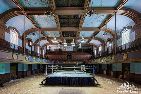 An abandoned boxing ring sits in a large arena with peeling paint and ornate architecture. A large audience area overlooks the ring.