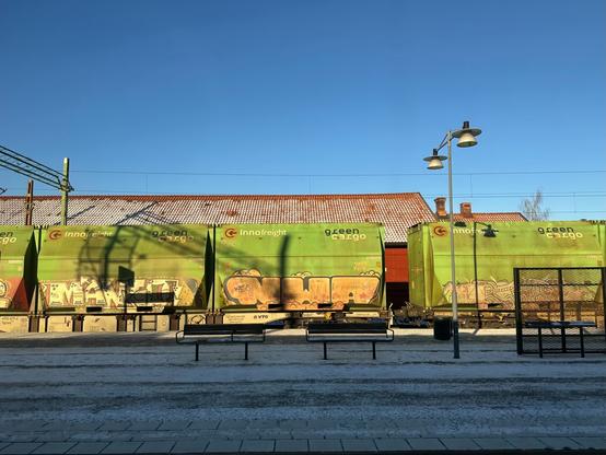 A line of green railway freight wagons, covered with faded graffiti lettering, passing through a snow-covered railway station.