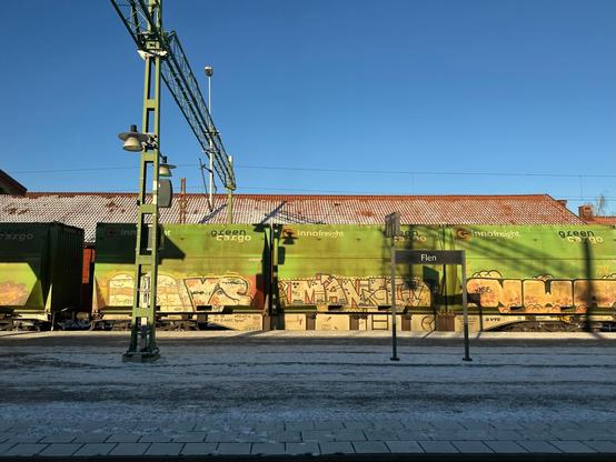 line of green railway freight wagons, covered with faded graffiti lettering, passing through a snow-covered railway station. A name board on the platform identifies the station as “Flen”.