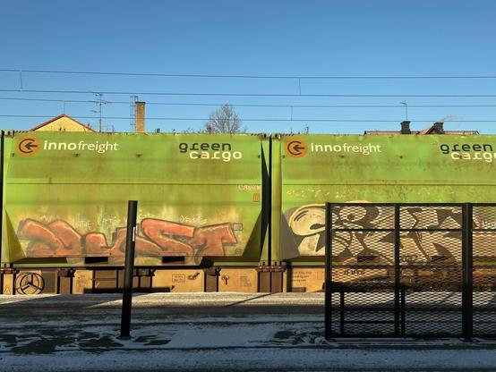 line of green railway freight wagons, covered with faded graffiti lettering, passing through a snow-covered railway station. A metal grille partly obscures the rightmost wagon and the piece on it