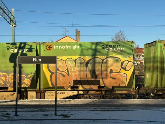line of green railway freight wagons, covered with faded graffiti lettering, passing through a snow-covered railway station. A name board on the platform reads “Flen”