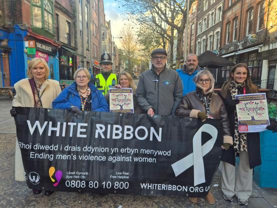 Group of people holding a White Ribbon banner and posters on a Swansea street, including a police officer and campaigners promoting domestic abuse awareness.