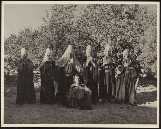 The image is a black and white photograph featuring nine individuals in what appears to be traditional or historical attire. They are standing outdoors with trees and bushes visible in the background, indicating an outdoor setting.

Starting from left to right: The first three people are wearing dark robes with head coverings that resemble nun's habits or headdresses commonly associated with certain religious orders. These figures have a solemn expression on their faces as they look directly at the camera. Following them is another individual dressed in darker clothing, possibly representing an older person given the appearance of grey hair and wrinkles.

In the center sits a younger figure wearing striped garments that could be indicative of traditional or ceremonial attire for boys during this period. This child holds what seems to be a round mirror on his lap while looking at it thoughtfully with one hand raised near their face as if in prayerful contemplation.

Behind these central figures stand six other individuals, all adorned in long robes and head coverings similar to those of the first three people but without any distinct markings or insignia. Their expressions vary from neutral to slightly joyful, suggesting they are partaking in a communal activity such as dancing at a wedding ceremony.

The photograph carries an air of authenticity and may have been taken during a historical reenactment event given its comp [...]
