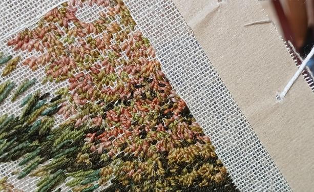 The top right hand corner of an unfinished stitched artwork. Lots if small red and green leaves. The taped edge of the canvas and lacing thread can be seen to the right