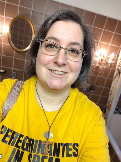 Spiegel-Selfie im Bad, mit glitzerndem Lidschatten, liquid Eyeliner und Wimperntusche. Ich trage ein sonnengelbes Vatikan-Shirt, mit dem lat. Motto des Hl. Jahres 2025 (Pilger*innen der Hoffnung). Dazu trage ich den üblichen Taukreuzanhänger mit kath. Medaillen, Brille und Handtasche an der re. Schulter sitzen auch. Ich lächle. Im Hintergrund die braunen Fliesen und goldene Dekoakzente.