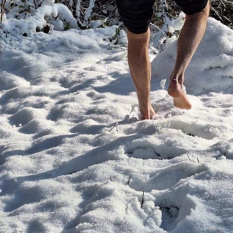 Zwei unbekleidete Füße gehen im Schnee davon. Über dem Knie beginnt die zusammengeraffte schwarze Hose. Am oberen Bildrand dunkles Gehölz. Die Sonne scheint und wirft lange Schatten nach links unten. Durch die Schneedecke ragen einzelne Grashalme.
