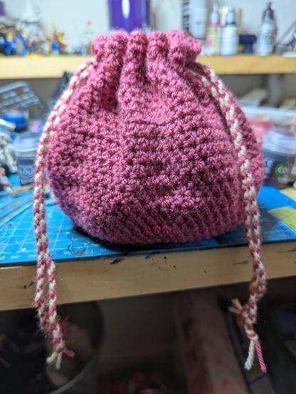 A raspberry pink crocheted dice bag, with pink and cream striped drawstrings pulling the top closed