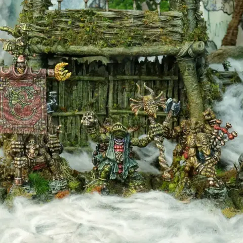 A group of intricately detailed miniatures inspired by Celtic mythology is displayed in front of a rustic, weathered structure covered in moss and foliage. The miniatures, depicting various fantastical creatures, include a central figure holding a banner, flanked by others wielding weapons and adorned with unique features like tusks and intricate patterns. The scene is set amidst a misty, ethereal atmosphere, enhancing the mystical theme of the Famhairean campaign.