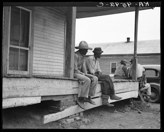 The black and white photograph depicts three men sitting on a wooden porch bench in front of what appears to be an old-fashioned rural house or barn. The man on the far left is wearing a straw hat, while the other two are wearing hats as well. They all seem relaxed, with their legs crossed or resting comfortably against one another. To the right side of the image, there's a glimpse of another person sitting down on what looks like a barrel near a vehicle, possibly engaged in some work-related activity.

The photograph has text at the top corner that reads "9-SPEC-AJ - AR," which could be indicative of its origin or cataloging system. The setting suggests an agricultural community with elements such as farming tools and vehicles present. This image captures everyday life in rural America during a specific historical period, likely associated with efforts to document social conditions for the Farm Security Administration (FSA) under President Franklin D. Roosevelt's New Deal programs.

The photograph carries significant documentary value due to its association with famous photographer Dorothea Lange, known for her work capturing American history through images of rural life and human experiences during times like The Great Depression and World War II. This particular image falls into the genre of social realism photography that Lange was renowned for.