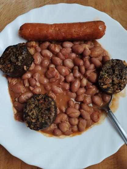 Una vista cenital de un plato blanco que contiene un guiso de judías en salsa rojiza. En la parte superior del plato, hay un chorizo grande y rojizo. Alrededor de las judías, se aprecian tres rodajas de morcilla de color oscuro y textura granulada. Una cuchara de metal plateado se asoma por el lado derecho del plato, que descansa sobre una superficie de madera clara.