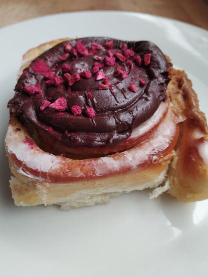 Un apetitoso cinnamon roll dulce, cubierto con un espeso glaseado de chocolate oscuro y espolvoreado con pequeños trozos de frambuesa liofilizada de color rosa vibrante. En los bordes del rollo se ve un glaseado blanco o rosado pálido. El cinnamon roll, de textura suave y esponjosa, descansa sobre un plato blanco.