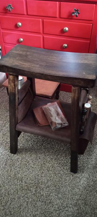 A small, simple, backless, medium brown coloured bench transformed into a work bench with panels containing leather working tools on either side and a small shelf between the legs under the work top. All the panels are made from thick medium brown waxed leather. It is on a mauve carpet. A red dresser peaks out from the background.