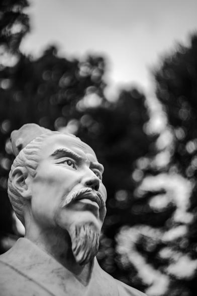 The image portrays a black and white close-up of a stone statue of a man with Asian features. The statue depicts a historical figure with a stern expression, having pronounced cheekbones and wearing a traditional topknot hairstyle. The figure has an intricately carved mustache and goatee. The background is softly focused, featuring blurred outlines of trees, giving a sense of depth and an outdoor setting.