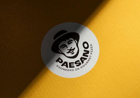 A close-up of a round logo on a yellow surface, featuring the text "PAESANO" and "Pioneers in Gourmet Pizza."