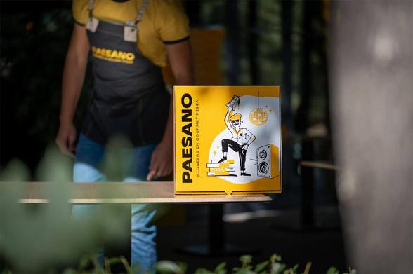 A pizza box labeled "PAESANO" featuring a playful illustration of a dancing chef, with a disco ball and speakers, in a vibrant outdoor setting.