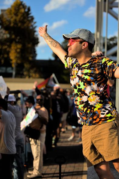 a man standing over a highway bridge with a crowd of protestors. he is on a barrier blocking the sidewalk from the road and hanging from a support pole blowing a kiss to the cars below.