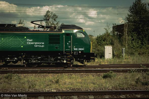 A modern, green locomotive waiting to depart; the words 'Rail Operations Group' and the numbers '93 004' are painted on its side. The railway tracks are surrounded by shrubs and small trees. The sky is blue and sunny with plenty of white clouds.