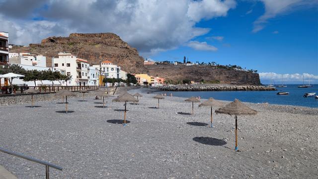 Una spiaggia di sassolini con ombrelloni di paglia e scogliere a ridosso delle case.