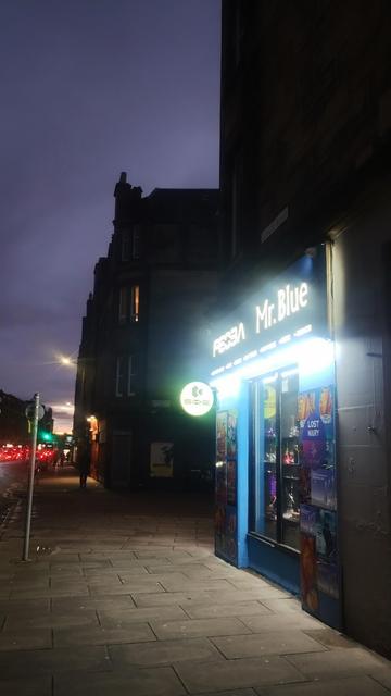 Looking along an urban street just after dusk, brightly lit, blue painted frontage of a shop in the foreground