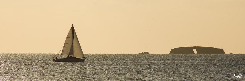 Photo panoramique montrant un paysage marin.
Sur le tiers gauche, un voilier, voiles déployées, se dirige vers la droite.
Sur le tiers droit, une île rocheuse presque rectangulaire dépasse sur l’horizon, et présente en son milieu un gros trou.
L’ambiance est un peu jaune révélant un ciel couvert et un éclairage en contre-jour, donnant au global une photo presque sepia.