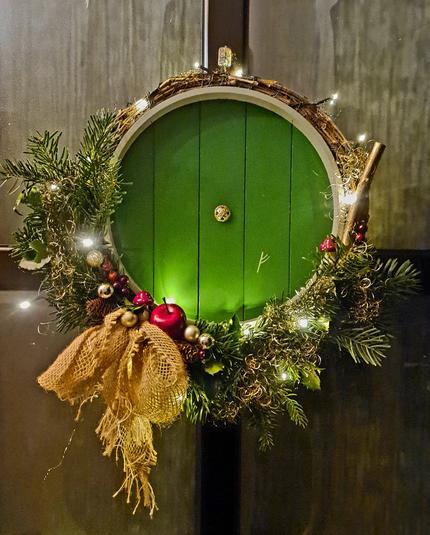 Türkranz mit hobbithöhlentür in der Mitte. Tür besteht aus einem runden Tablett, grün angemalt mit goldknopf in der Mitte und g-rune rechts unten. Um die Tür ist ein Kranz aus Ranken gewickelt, asymmetrisch mit Tannen und stechpalme. Deko sind fliegenpilze, goldene Kugeln, ein Apfel und eine große Schleife aus Rupfen.