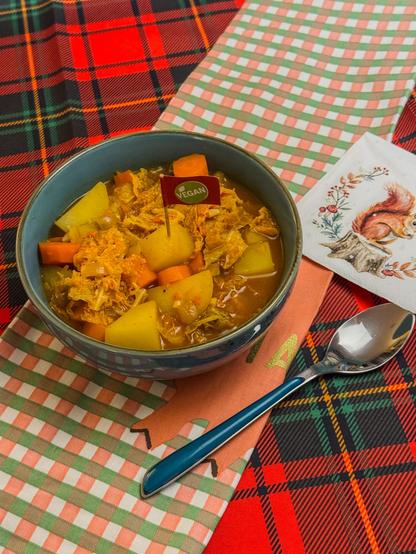 Eine Nahaufnahme zeigt eine blaue Schüssel mit einem Eintopf auf einem karierten Tuch. Der Eintopf enthält geschnittene Karotten, Kartoffeln und Kohl. Eine blauerEsslöffel liegt in der Nähe der Schüssel. Die Schüssel steht auf einem quadratischen Stofftuch in Rot-, Grün- und Weißtönen. Auf der rechten Seite befindet sich eine quadratische Unterlage mit einem Motiv von Eichhörnchen. Auf der Schüssel ist ein kleines, rundes Etikett mit der Aufschrift "VEGAN" zu sehen.