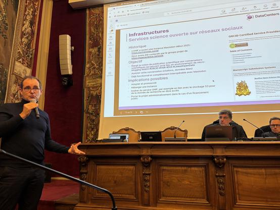 Photo prise dans la salle du Sénat de l’université Paris Cité, reconnaissable à ses tentures. Je tiens le micro à gauche et sur la droite se trouvent à La Tribune le directeur de l’UNIF, Dominique Bascle et Gilles Roussel président de son assemblée général. L’écran au dessus d’eux affiche une page de présentation où l’on reconnaît en bas à droite le logo de bonfire.