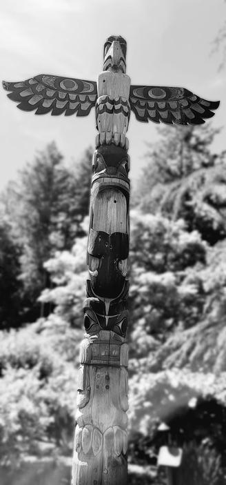 Black and white photograph of a large, carved wooden totem pole, depicting various stylized figures and an eagle with outstretched wings at the top, against a blurred background of trees.