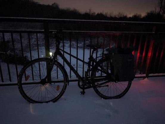 Ein Fahrrad mit leuchtendem Vorder- und Rücklicht lehnt an einem Brückengeländer. Es ist dunkel und es liegt Schnee.