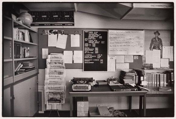The image depicts a vintage newsroom office filled with various objects that provide insight into the activities and work environment of journalists during this period. In the center, there's a large corkboard plastered with papers, schedules, maps labeled "Tokio," "Moskau," "Washington," and more indicating different cities or regions for which correspondents report. A handwritten blackboard shows various news items such as crime rates in Los Angeles and London. The board is cluttered but organized, indicative of a busy reporting environment.

A typewriter sits on the desk to the right, symbolizing traditional journalism before digital media took over. Above it are shelves stocked with books and folders labeled "Amsterdam," suggesting extensive research materials for journalists covering global events. A globe in front of the bookshelf underscores the international scope of news coverage.

On the left side, a stack of newspapers such as 'Die Welt,' 'De Standaard,' and others from various European countries are visible, indicating sources used by these reporters or editors. The overall atmosphere is one of intense focus on delivering timely information to readers around the world, with an air of historical significance given its monochromatic nature and dated objects like typewriters.

This photo captures a slice of journalistic history at a time when news was disseminated manually through print media across continents.  [...]