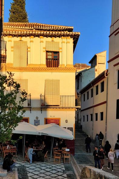 Los últimos rayos de sol se reflejan en una fachada de la cuesta de San Gregorio. En la parte de abajo, en el bar el 22 hay gente sentada en la terraza, debajo de las sombrillas. Alguna gente camina cuesta arriba por la calle.