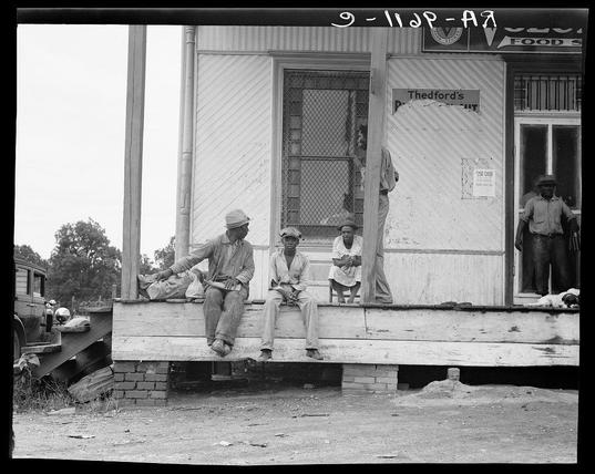 This black and white photo captures a moment in time at what appears to be an African American community, possibly on or near a plantation. Four men are featured prominently; three seated with their backs against the wooden porch of a weathered structure labeled "Thedford's" above its entrance, which indicates it could serve as both a store and perhaps lodging or dining establishment based on signage that reads "Food S." A fourth man stands in the background near another sign offering cash. The men are dressed in work clothes typical for rural laborers; two wear hats while all four don overalls and simple shoes suitable for fieldwork.

The porch is elevated, supported by a series of wooden beams with some showing signs of deterioration or repair attempts through nailed patches. A brick wall underlines the structure's foundation. To the left side in front of this rustic building sits an old truck, which suggests transportation methods from that era were simple and utilitarian.

A sense of camaraderie is evident among these individuals as they sit together on a bench built to fit their needs; one man reclines comfortably with his legs crossed while another rests both hands on what looks like a bag or sack beside him. Their relaxed postures indicate the photo may have been taken during a break from work.

The scene exudes an aura of resilience and community spirit, as these men find rest in each other's company amidst their [...]