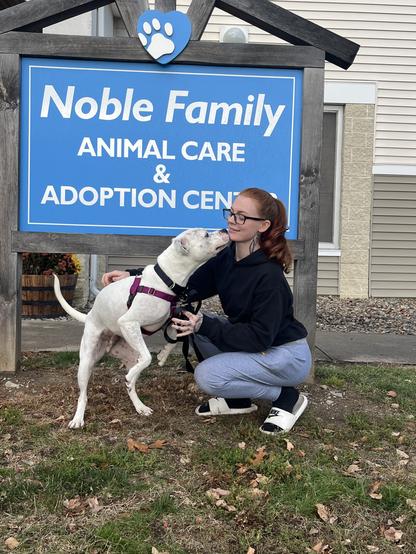 Ultra, a pittie mix, stands outside the nevins farm shelter with his new owner. ultra is medium sized and mostly white with some faint black spots. he's licking the woman's face as she crouches next to him. she has long red hair in a ponytail and is wearing a dark top with light jeans. they're in front of a sign that says noble family animal care and adoption center.