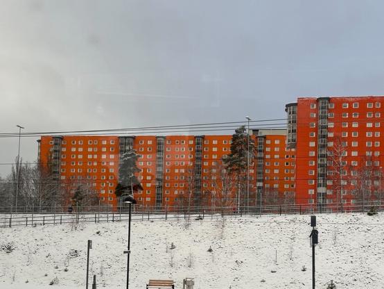 Two large orange-red apartment blocks looming over a snow-covered embankment. In front of it, a railway station platform can be made out