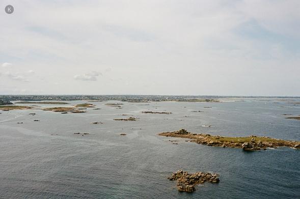 Weite Meereslandschaft unter hellem, wolkigem Himmel; ruhiges Wasser mit verstreuten kleinen Felsen und Inseln, ferner Küstenstreifen am Horizont.