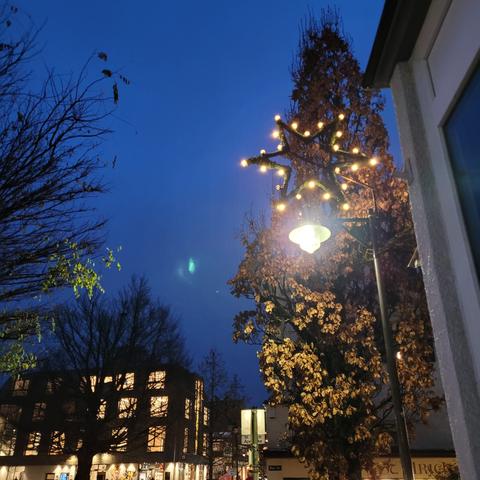 Szene in der Staft beim Staftplstz. Rechts Häuser, eine Laterne darüber ein großer leuchtender Stern. Davor ein Baum der im Licht leuchtet. Links ein großes neues Gebäude mit vielen Fenstern die hell erleuchtet sind.