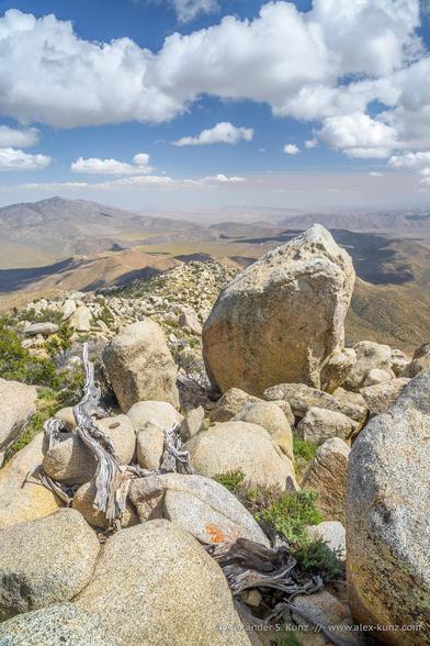 A vertical orientation photo showing a desert landscape. In the immediate foreground are big granitic rocks, in the distance valleys and barren desert mountains. Clouds cast their shadows onto the land.