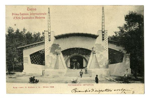 The image depicts a historical black and white photograph of the entrance to Torino's First International Exhibition of Modern Decorative Art. The building has an archway with the text "Fotografia Artistica" prominently displayed on it, indicating its function as an art photography exhibition space within this larger event. Two large, ornate towers flank both sides of the entryway, each featuring intricate designs and what appears to be a staircase leading up to them.

There are several people visible near the entrance: one walking towards the stairs with luggage in hand, another standing on top of the steps wearing a suit hat, possibly taking a photo or admiring the view. The setting suggests an outdoor environment as trees can be seen behind and beside the building. A photographer's credit is noted at the bottom left corner "Fotografia Artistica" by G. Modiano from Milan in 1936.

The upper portion of the image contains text, likely providing information about the event and its location: "Torino - Prima Esposizione Internazionale d'Arte Decorativa Moderna," which translates to Turin's First International Exhibition of Modern Decorative Art. There is also a signature or marking at the bottom right corner that appears handwritten in ink.

The photograph captures not only an architectural element from this historical event but also provides insight into fashion and social customs indicative of early 20th-century Italy, wi [...]