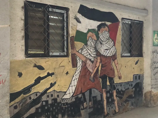 Graph sur un mur montrant deux enfants en keffieh avec un drapeau palestinien, et autour, des fusils pointés sur eux et des bombes tombant sur la ville en arrière plan.
