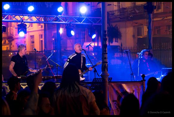 Live music performance photograph showing the band Big Generator performing on an outdoor stage with blue and purple LED lighting and overhead metal truss rig, featuring band members with guitars and vocals, a silhouetted audience in the foreground, and period architecture of Cobh visible in the background under evening light.