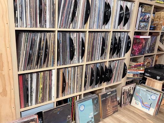 A view of the record shelf with dividers stuck in between letters of the alphabet.  New/uncleaned/uncatalogued records on the floor nearby leaning on the shelf