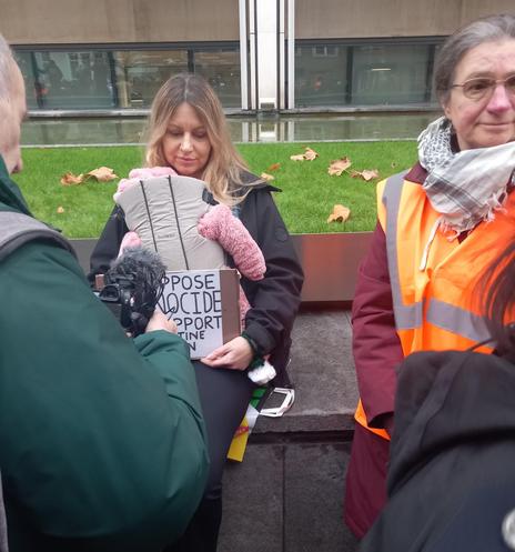 The image shows a woman holding a baby carrier in front of a glass building exterior. The womon is wearing a dark jacket and has shoulder-length light brown hair. They are holding a baby carrier against their chest, with the baby’s head visible. To the right of the woman, another person is wearing an orange safety vest and a red scarf. A sign held by the first person is visible with the text "OPPOSE GENOCIDE PORT". The ground is covered in fallen leaves,.