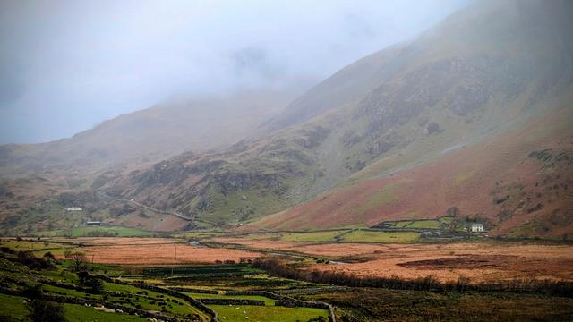 A misty valley landscape in Wales. The foreground features green fields and rugged stone walls enclosing sheep, leading into a wide expanse of rust-colored moorland and marsh. In the middle distance, a small white farmhouse with a dark roof sits nestled against the base of a steep, grassy mountain slope. The background is dominated by large, imposing hillsides, their peaks obscured by a low-hanging, soft mist or fog. The overall mood is dramatic and rugged.