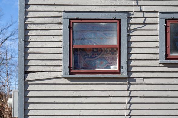 A window with a decorative curtain containing reds, blues and teals which complement the window exterior colors. A bit of siding is hanging down breaking the uniformity of the wall.