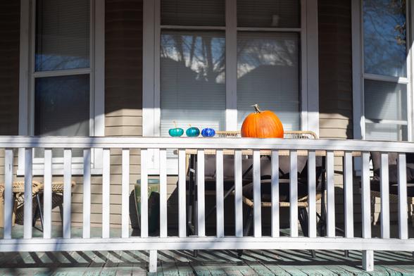 Teal decorative fake pumpkins with a real pumpkin beside them sit on a porch with a visible fence in the foreground.
