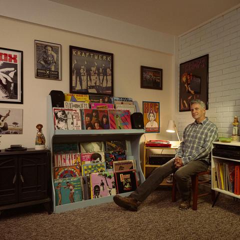 A white man with grey hair sits in an easy chair with his legs stretched out in front of him. He wears grey flannel and khaki work pants. He is surrounded by a collection of vintage country and punk LPs and posters.